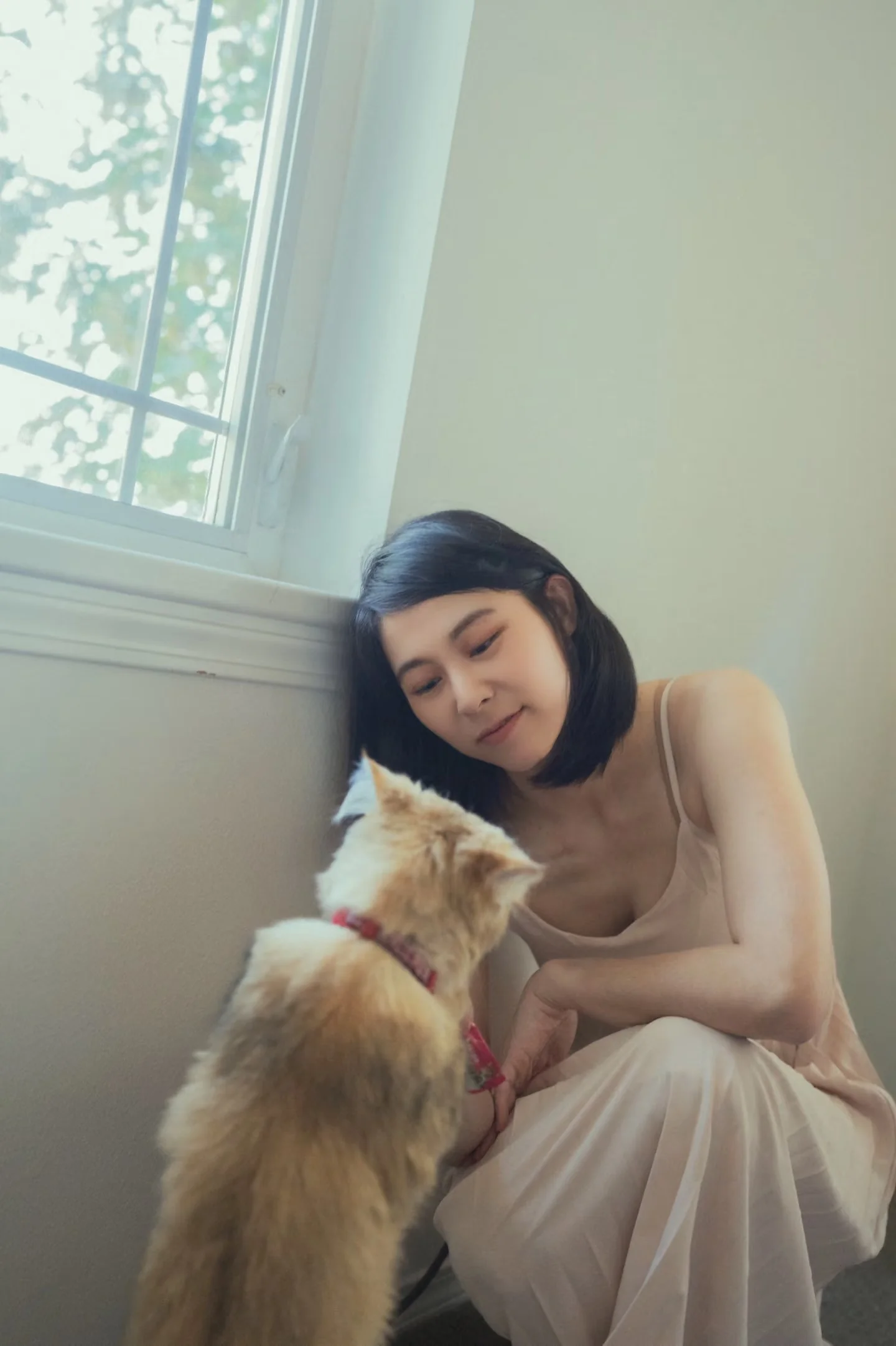 A woman and an orange cat facing one another beside a bedroom window