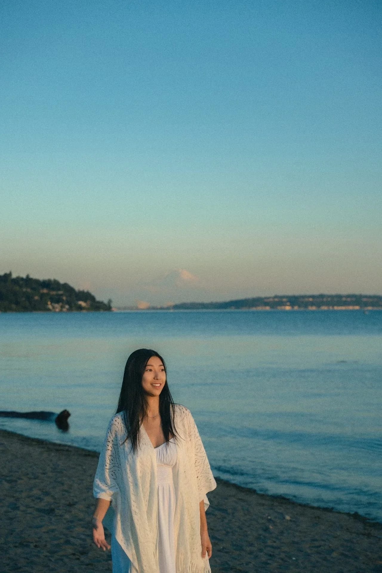 A woman standing beside calm water under a pale blue sky