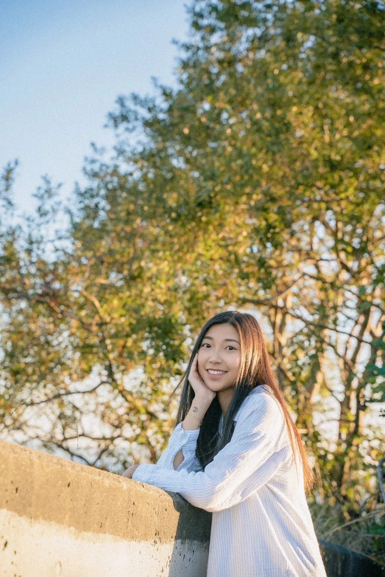 A smiling woman leaning on a low wall beneath sunlit trees