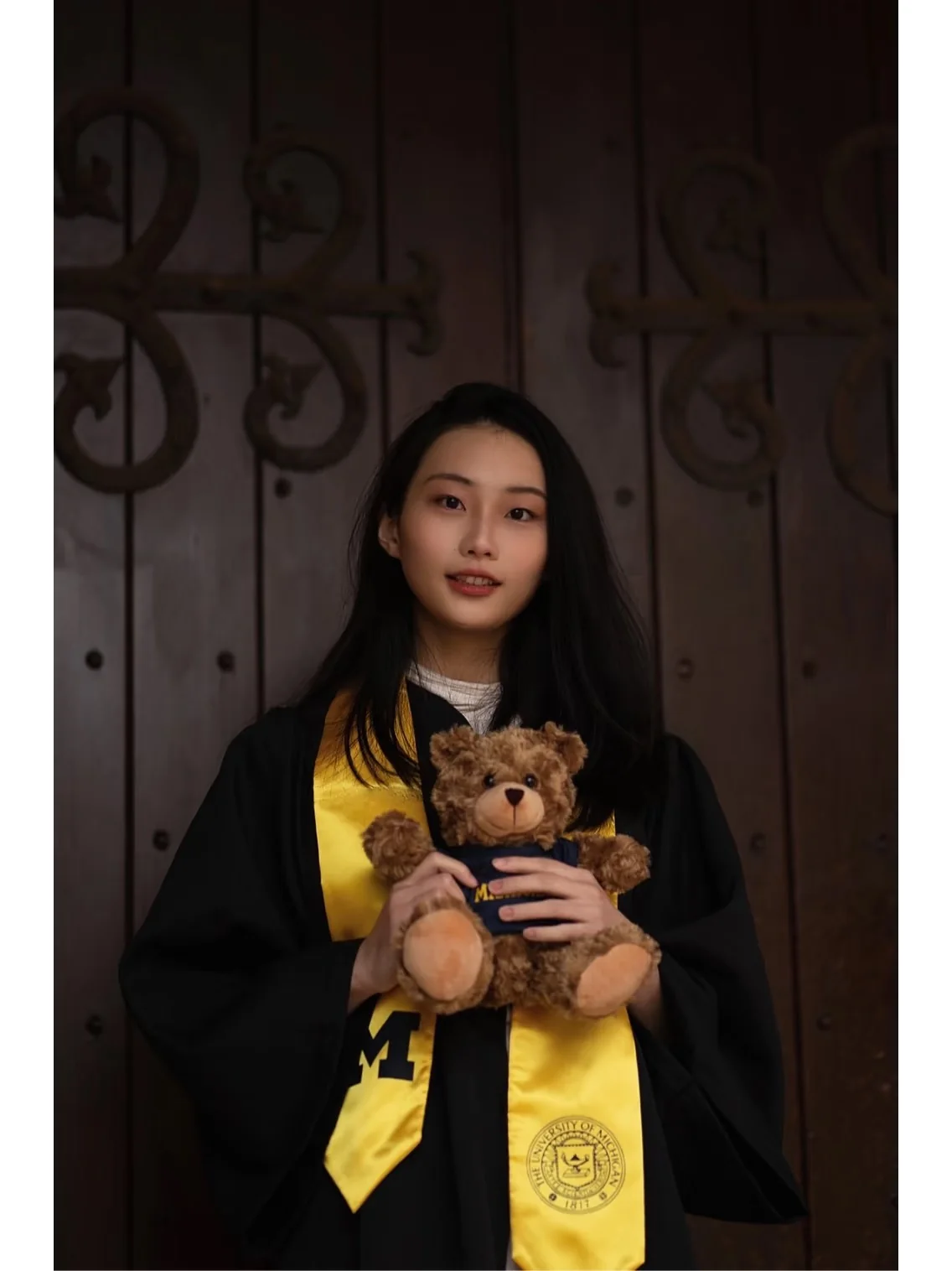A graduate in a black gown holding a teddy bear at a wooden doorway