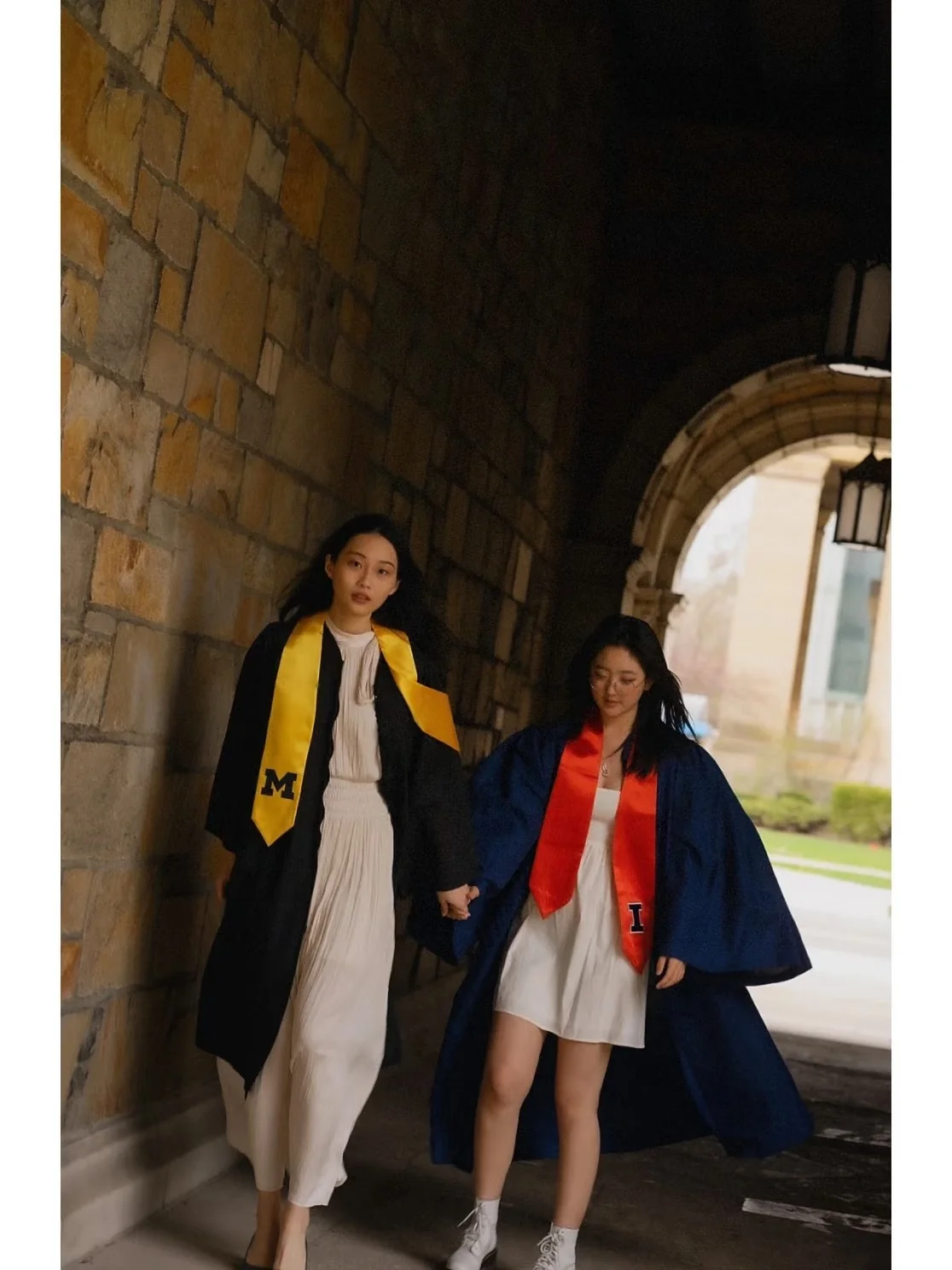 Two graduates walking together through a stone campus passage