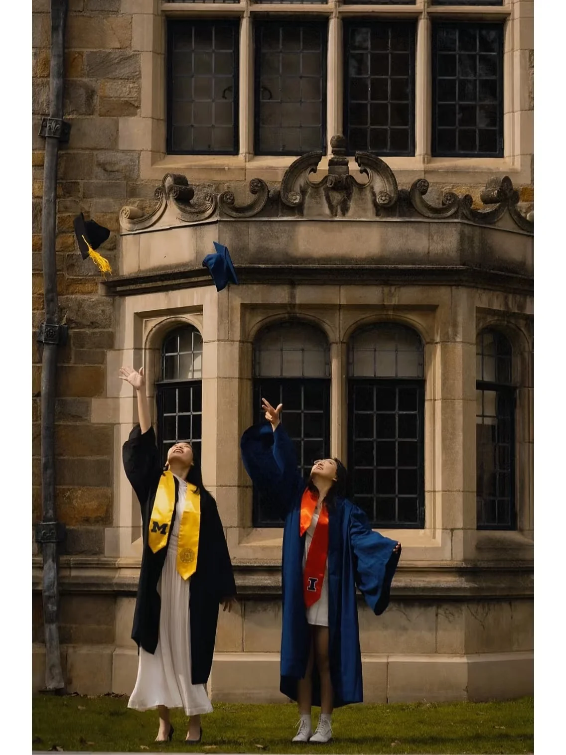Two graduates tossing their mortarboards in front of a stone campus building