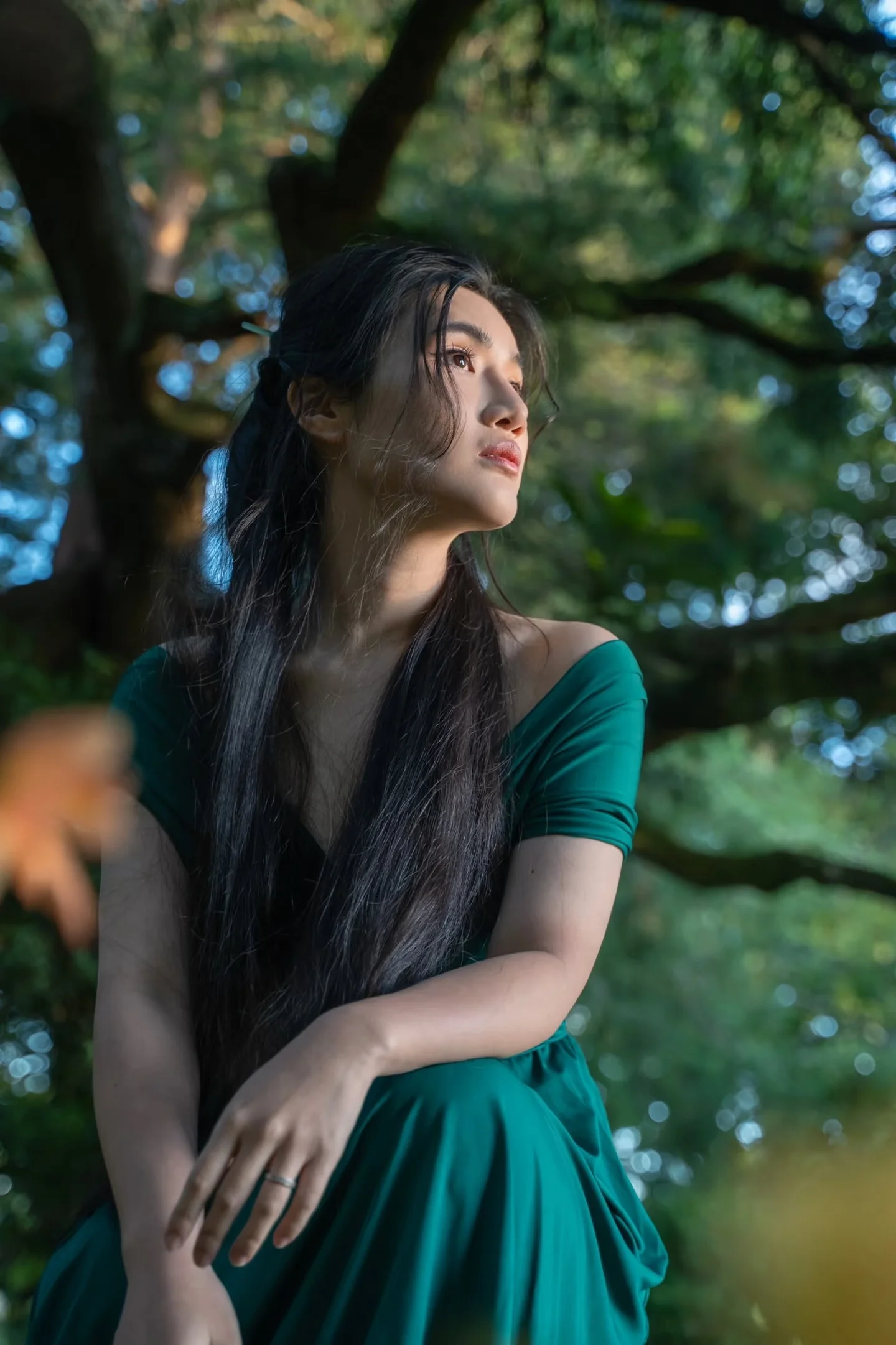 A woman in a green dress looking upward beneath leafy trees