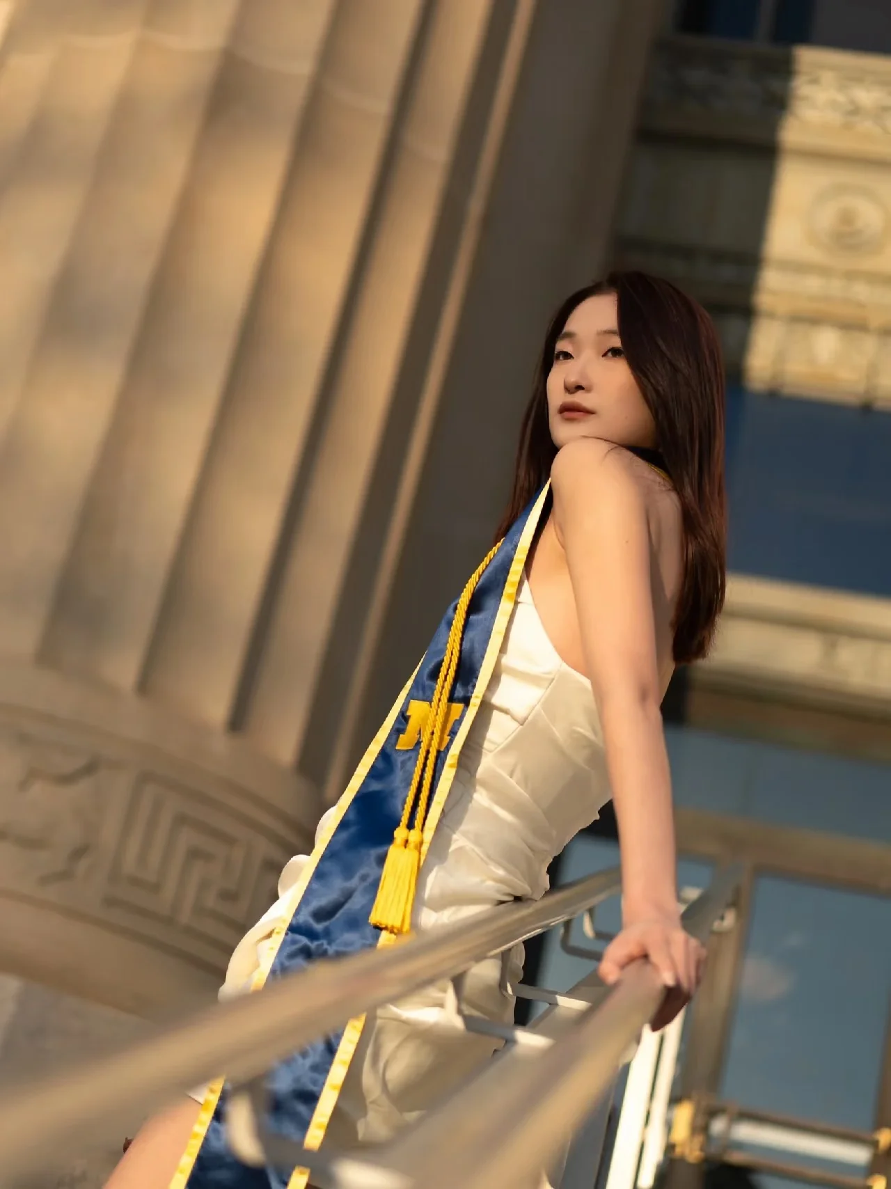 A graduate looking back from a railing beneath a campus colonnade