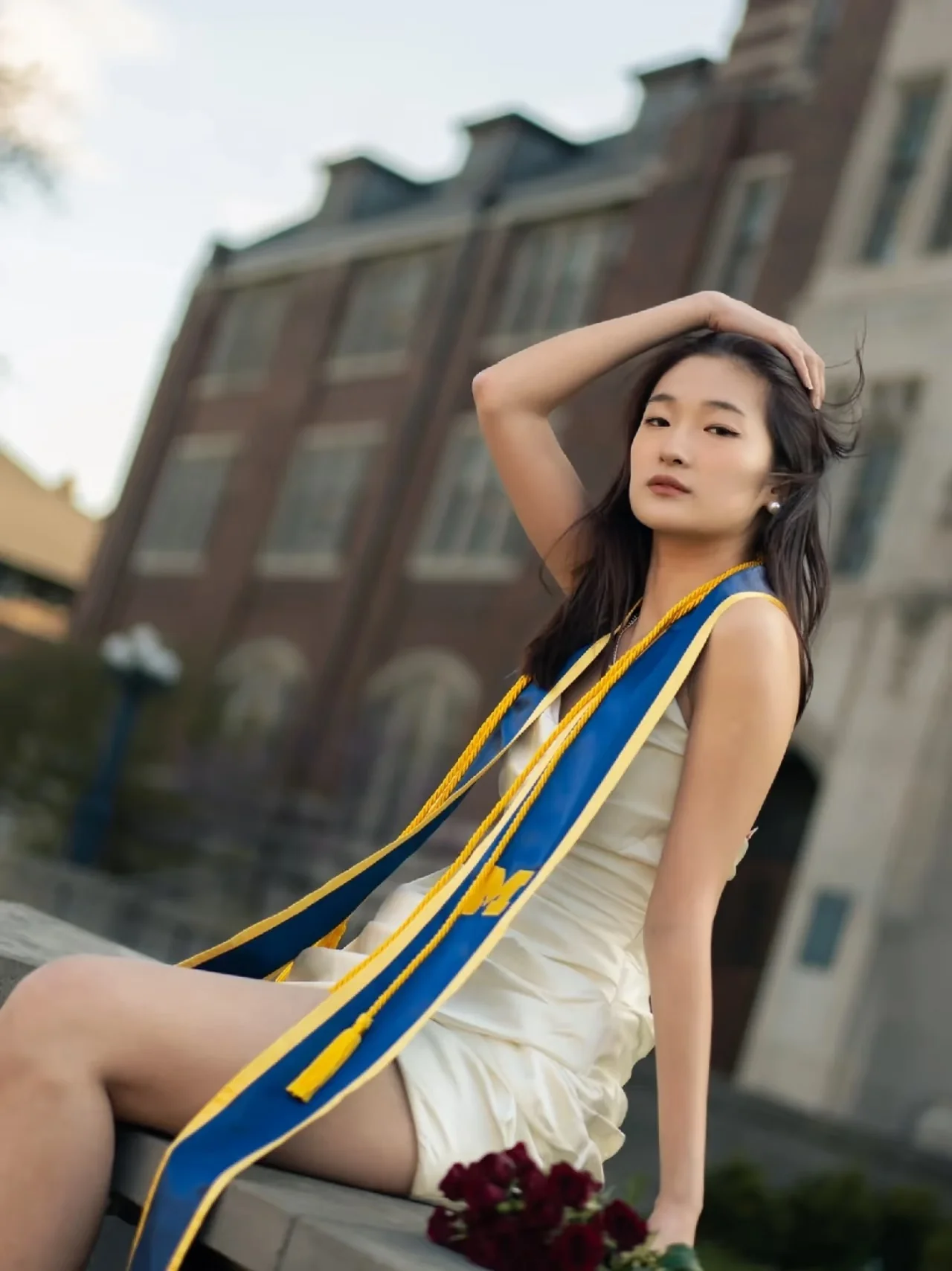 A graduate in a white dress and blue-and-yellow sash seated beside dark flowers