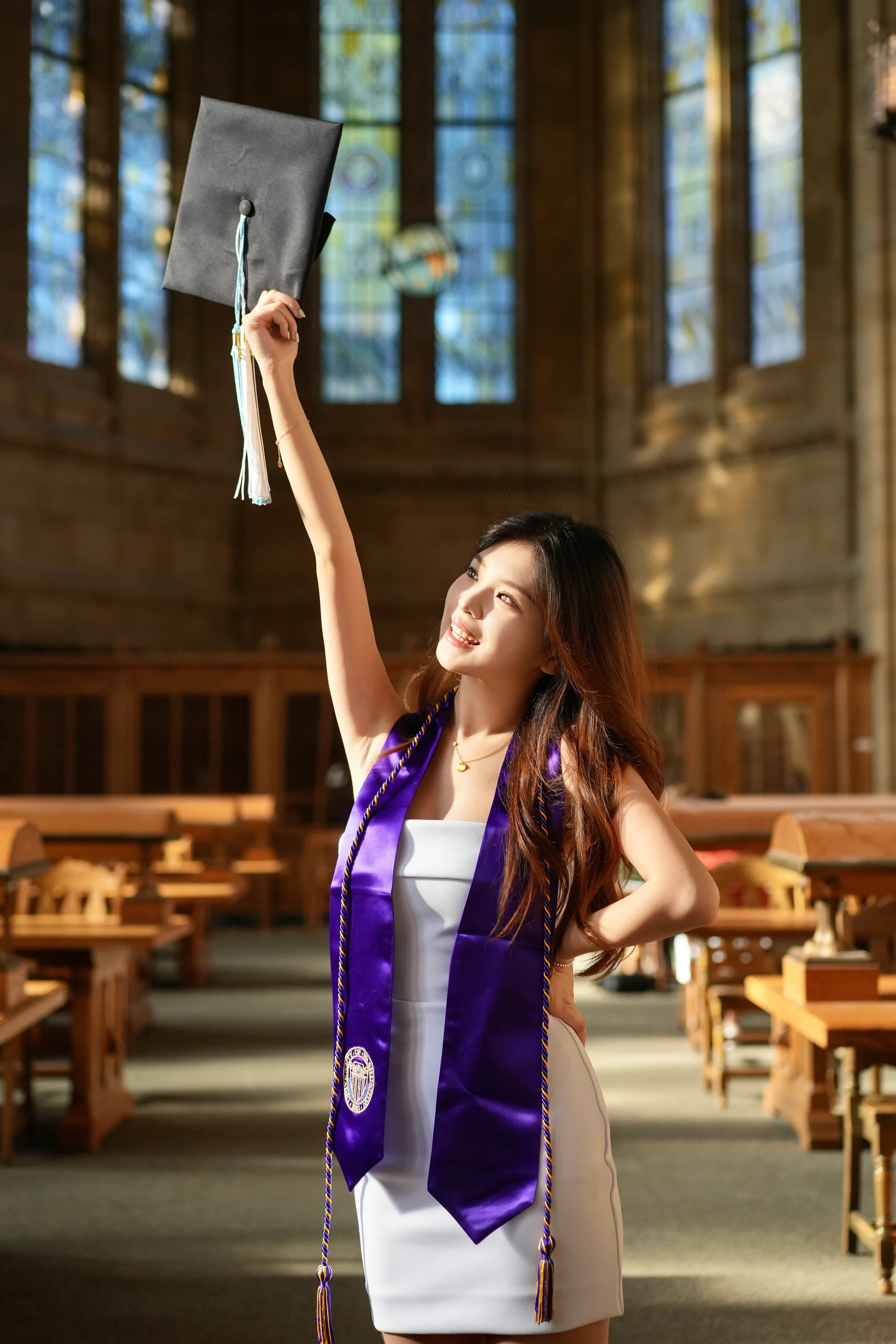 A graduate lifting a mortarboard in a sunlit reading room