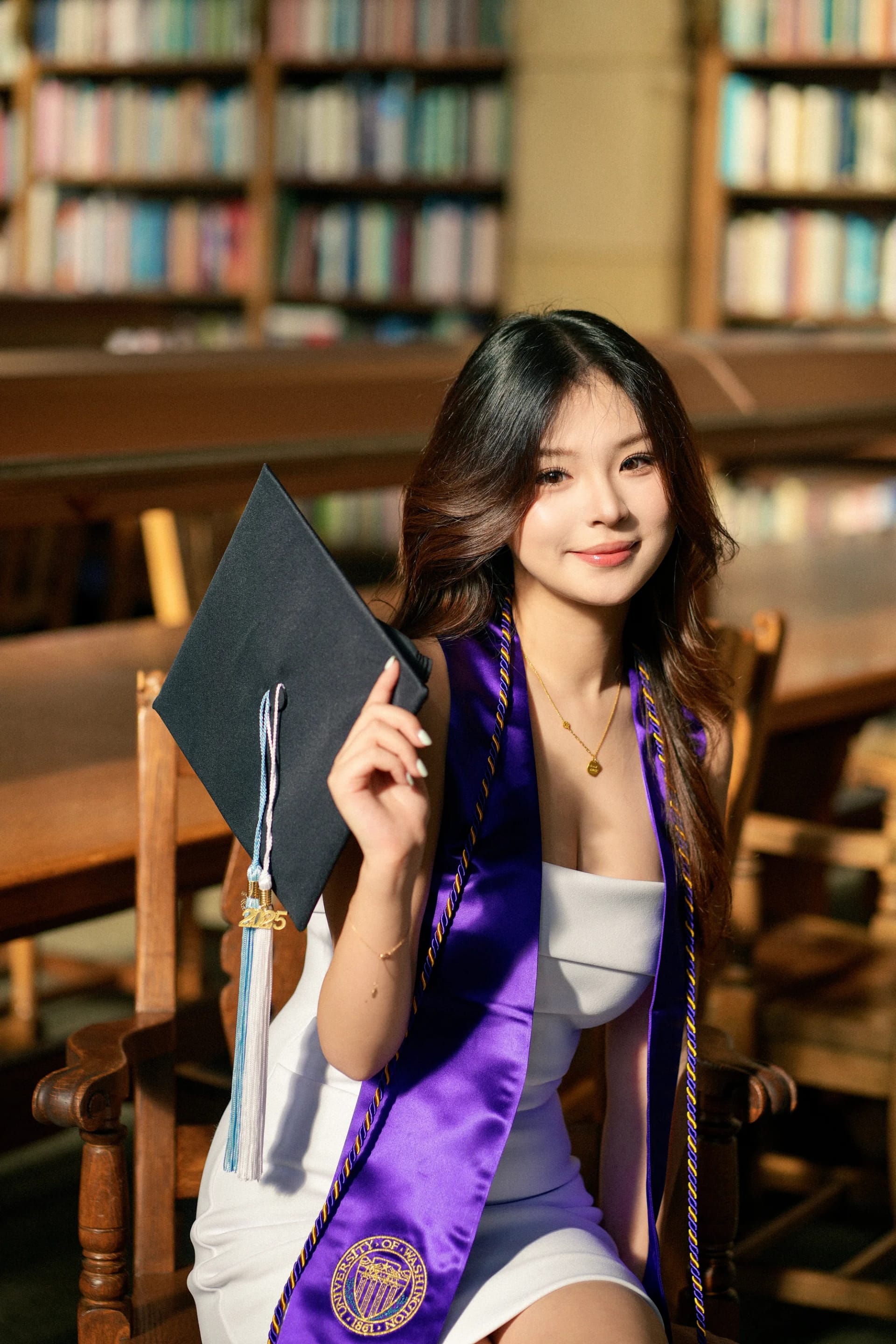 A graduate holding a mortarboard beside library bookshelves